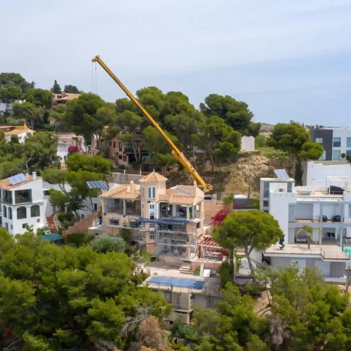 Fotografía aérea frontal de una casa unifamiliar en proceso de reforma con grúa de gran tonelaje entre pinos.